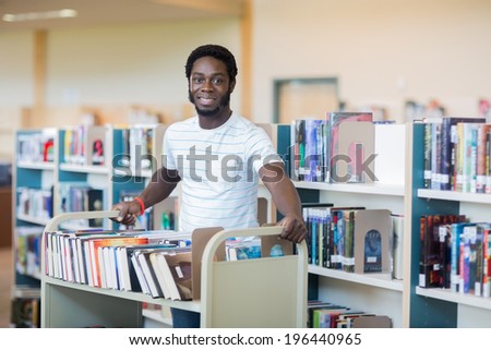 Portrait of young male librarian with trolley of books in library