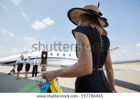 Rear view of rich woman with shopping bags walking towards private jet while pilot and stewardesses standing at airport terminal Rear view of rich woman with shopping bags walking towards private jet while pilot and stewardesses standing at airport terminal