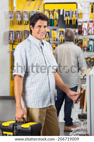 Portrait of smiling male customer purchasing tools with man in background at hardware shop