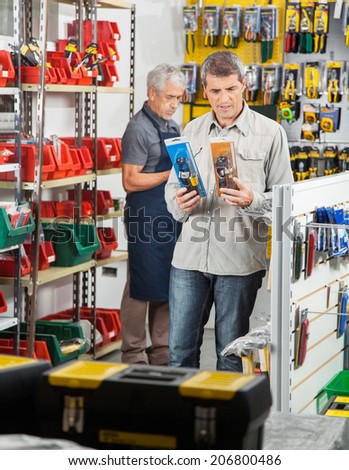 Mature man choosing soldering iron with worker working in background at hardware store