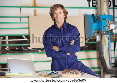 Portrait of confident carpenter standing with arms crossed against vertical panel saw
