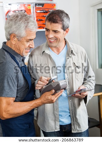 Senior man holding electronic reader while male customer paying through smartphone in hardware store