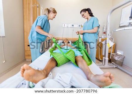 Nurses preparing male patient before transferring him on sling lift on bed at hospital
