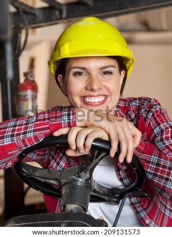 Portrait of happy female engineer wearing hardhat while leaning on steering wheel of forklift in workshop