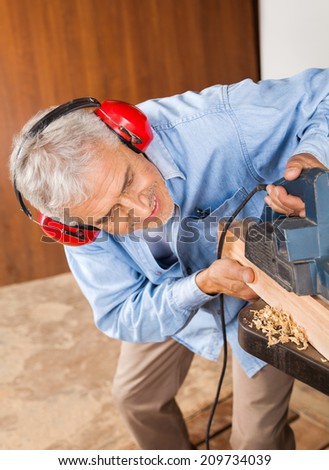 Senior carpenter wearing ear protectors while shaving wood with electric planer in workshop