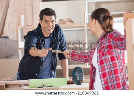 Happy young carpenter receiving hammer from female colleague in workshop