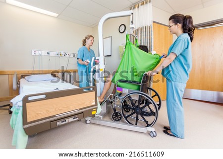 Female nurses transferring male patient from hydraulic lift to wheelchair in hospital