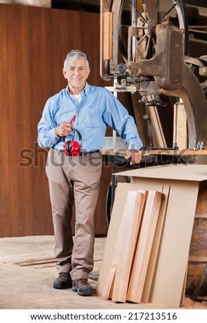 Full length portrait of senior male carpenter holding ear protectors by bandsaw in workshop