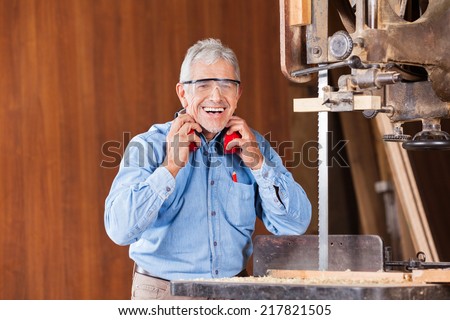 Portrait of happy senior male carpenter holding ear protectors by bandsaw in workshop
