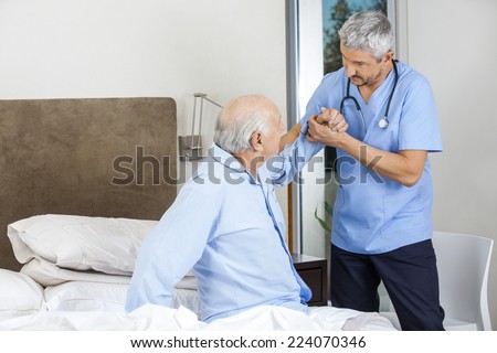 Male caretaker assisting senior man to get up from bed at nursing home