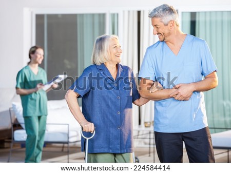 Happy male nurse assisting senior woman to walk with caretaker in background at nursing home yard