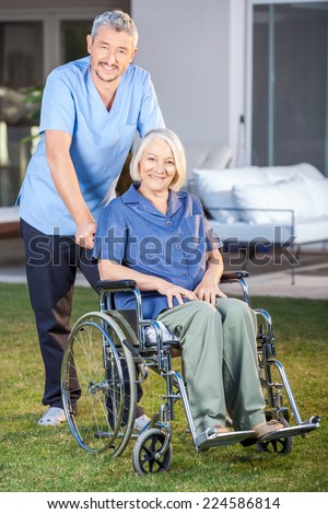 Full length portrait of male nurse standing with senior woman on wheelchair at nursing home lawn