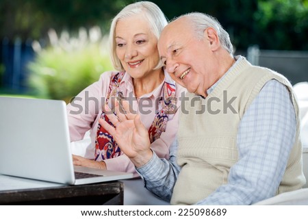 Happy elderly couple video chatting on laptop while sitting at nursing home porch