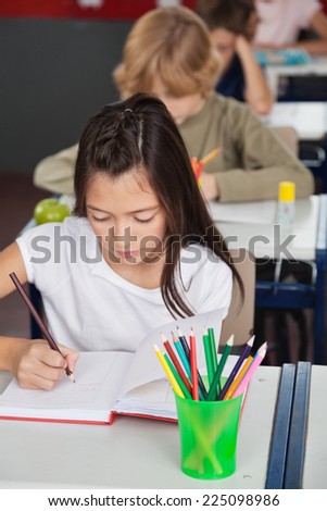 Little schoolgirl writing in book with classmates in background at classroom