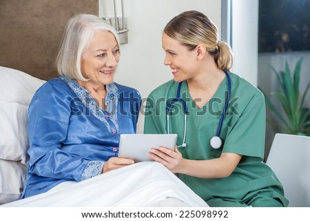 Happy female nurse and senior woman discussing while using tablet PC in bedroom at nursing home