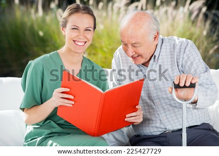 Portrait of happy female nurse reading book for senior man at nursing home