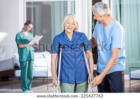 Male nurse helping senior woman to use crutches with caretaker in background at nursing home