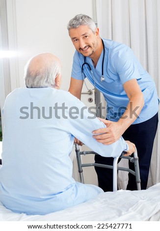 Portrait of smiling male caretaker helping senior man with walking frame in bedroom at nursing home