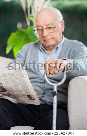 Elderly man reading newspaper while holding metal cane on couch at nursing home porch
