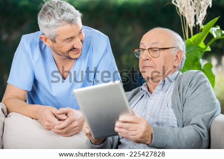 Smiling male caretaker looking at senior man using tablet computer at nursing home porch Smiling male caretaker looking at senior man using tablet computer at nursing home porch