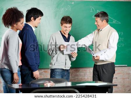 Happy male professor giving exam result to student with classmates standing in a row at classroom