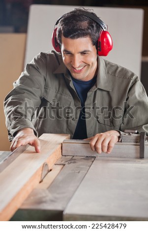 Happy young carpenter cutting wooden plank with tablesaw in workshop
