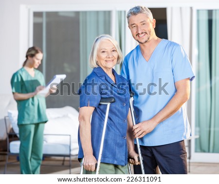 Portrait of happy male caretaker and disabled senior woman with female nurse in background at nursing home yard