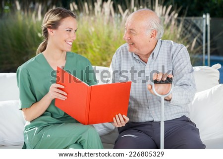 Smiling female nurse and senior man looking at each other while reading book at nursing home