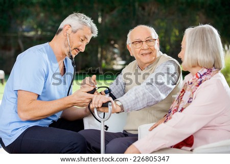 Mid adult doctor measuring blood pressure of senior man sitting beside woman at nursing home porch