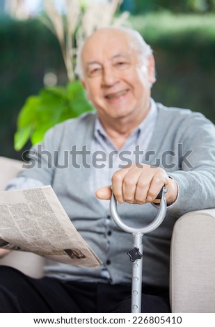 Portrait of happy senior man with newspaper and metal cane sitting on couch at nursing home porch