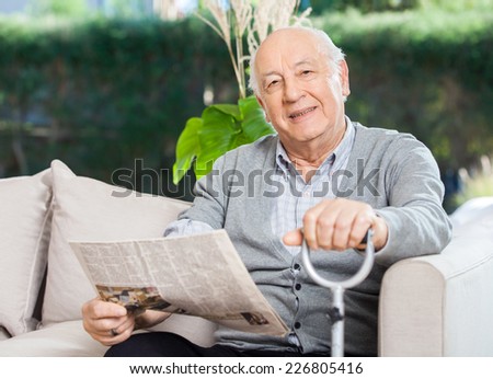 Portrait of confident senior man with newspaper and walking stick sitting on couch at nursing home porch