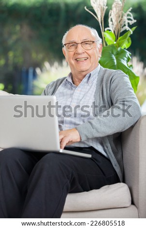 Portrait of happy senior man sitting with laptop on couch at nursing home porch