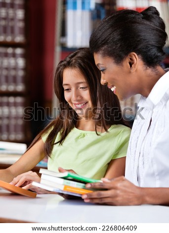 Happy female librarian and schoolgirl looking together at book in library