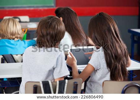 Rear view of little schoolgirl using digital tablet with boy at desk in classroom