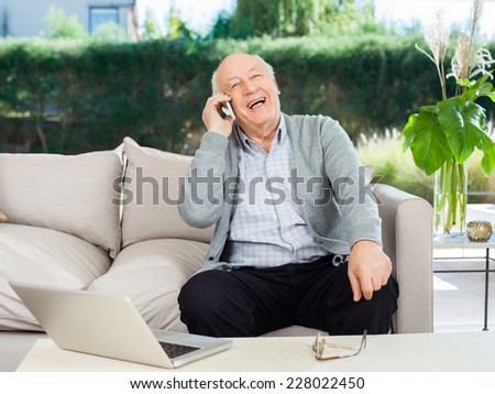 Cheerful senior man laughing while answering smartphone on couch at nursing home porch