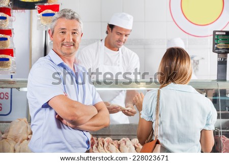 Portrait of confident mature man standing with arms crossed while woman choosing chicken meat in background at butcher\'s shop
