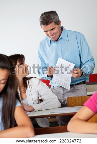 Happy male professor showing paper to student at desk during examination in classroom