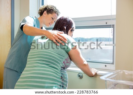 Mid adult female nurse comforting tensed pregnant woman leaning on window sill in hospital room
