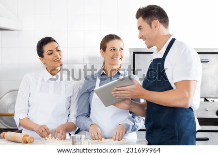 Happy chefs discussing recipe on digital tablet while preparing pasta in commercial kitchen