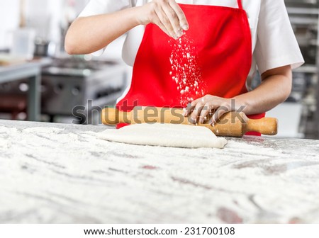 Midsection of female chef sprinkling flour while rolling dough at messy counter in commercial kitchen