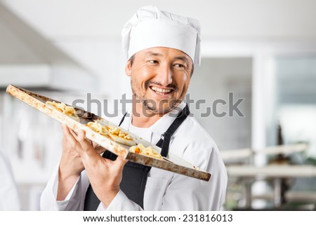 Happy male chef looking away while holding small pizzas on baking sheet in commercial kitchen