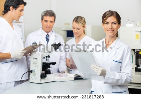 Portrait of female scientist holding clipboard with colleagues in background at laboratory