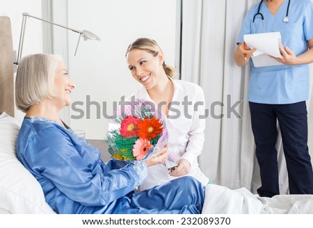 Happy daughter giving flower bouquet to sick mother at nursing home with caretaker in background