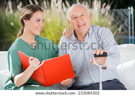 Portrait of happy senior man sitting by female nurse holding book at nursing home