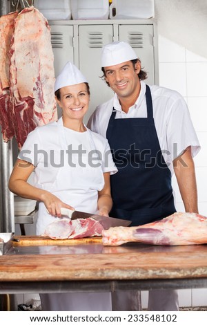 Portrait of confident female butcher cutting meat while standing by colleague at counter in shop
