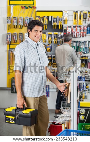 Portrait of confident young male customer buying hammer with man in background at hardware store