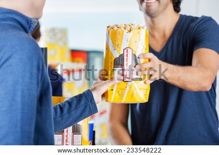 Midsection of male seller giving popcorn to man at cinema concession stand