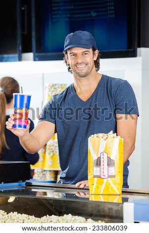 Portrait of happy male worker holding drink with popcorn at cinema concession stand while colleague working in background