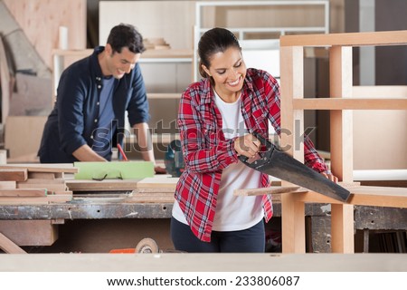 Happy female carpenter cutting wood with handsaw while colleague working in background at workshop