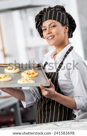 Portrait of confident female chef holding small pizzas on baking sheet in commercial kitchen